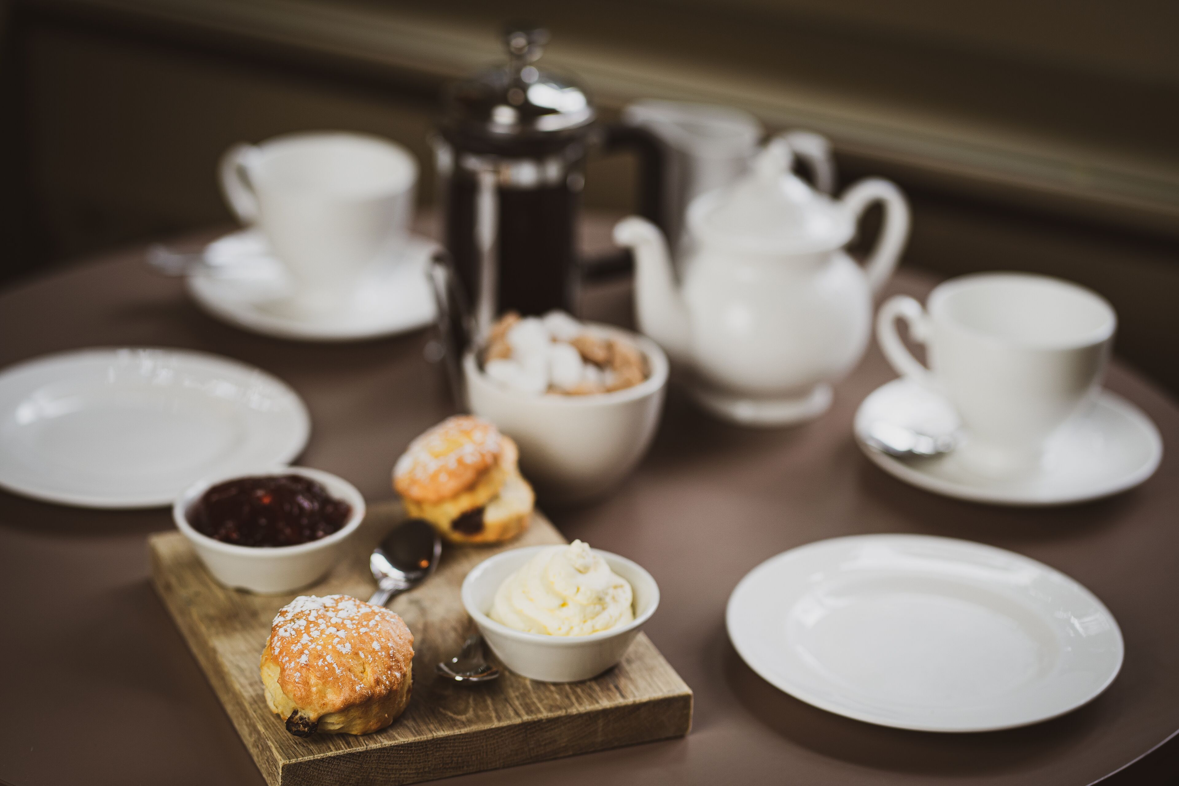 A table with a coffee pot, teapot, cups, saucers, a bowl of jam, a bowl of butter, and two scones on a wooden board.