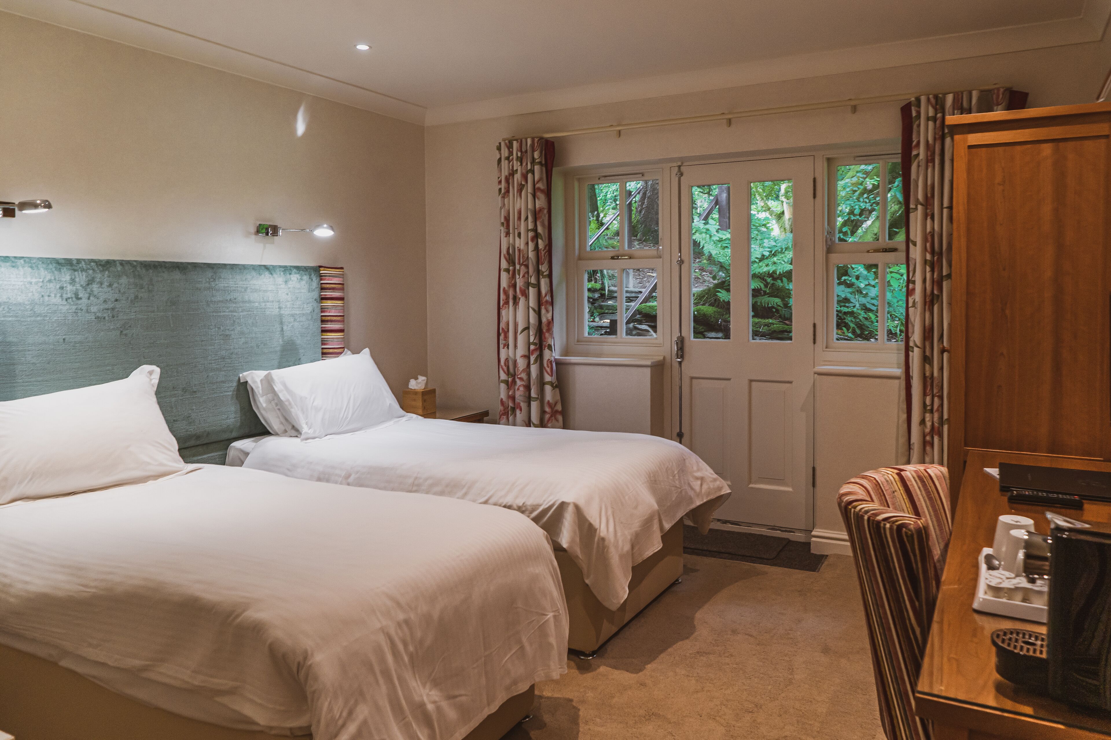 A hotel room with two beds, a wooden desk with a coffee maker, and a view of greenery through a window.