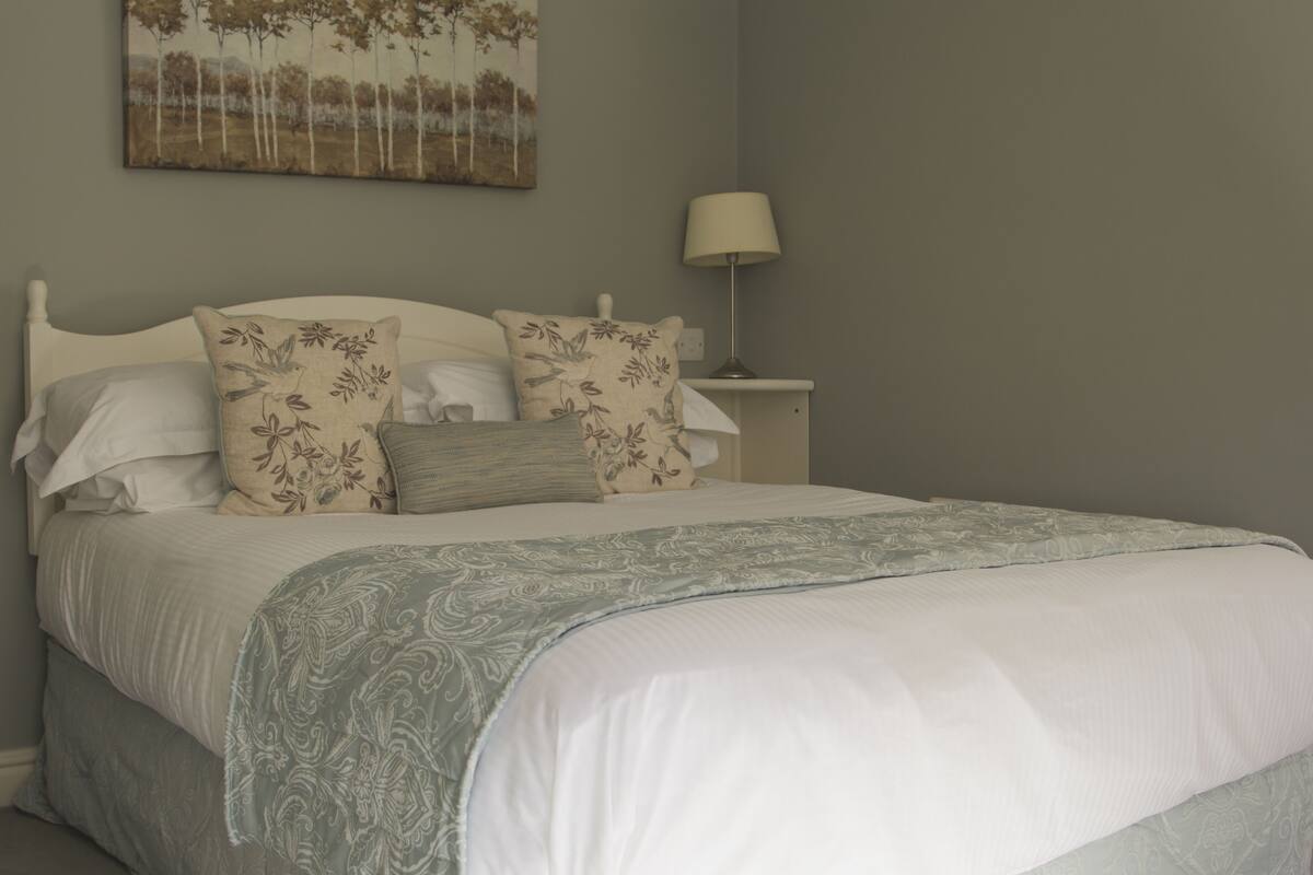Een opgemaakt bed met bloemenkussens, een bedlamp en een schilderij aan de muur.