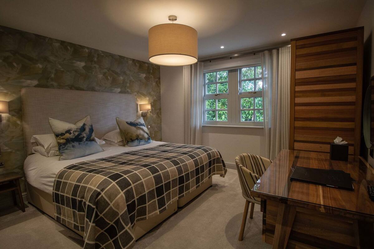 Een slaapkamer met een bed, nachtkastjes, een houten hoofdbord, een raam met gordijnen en een eettafel met stoelen.