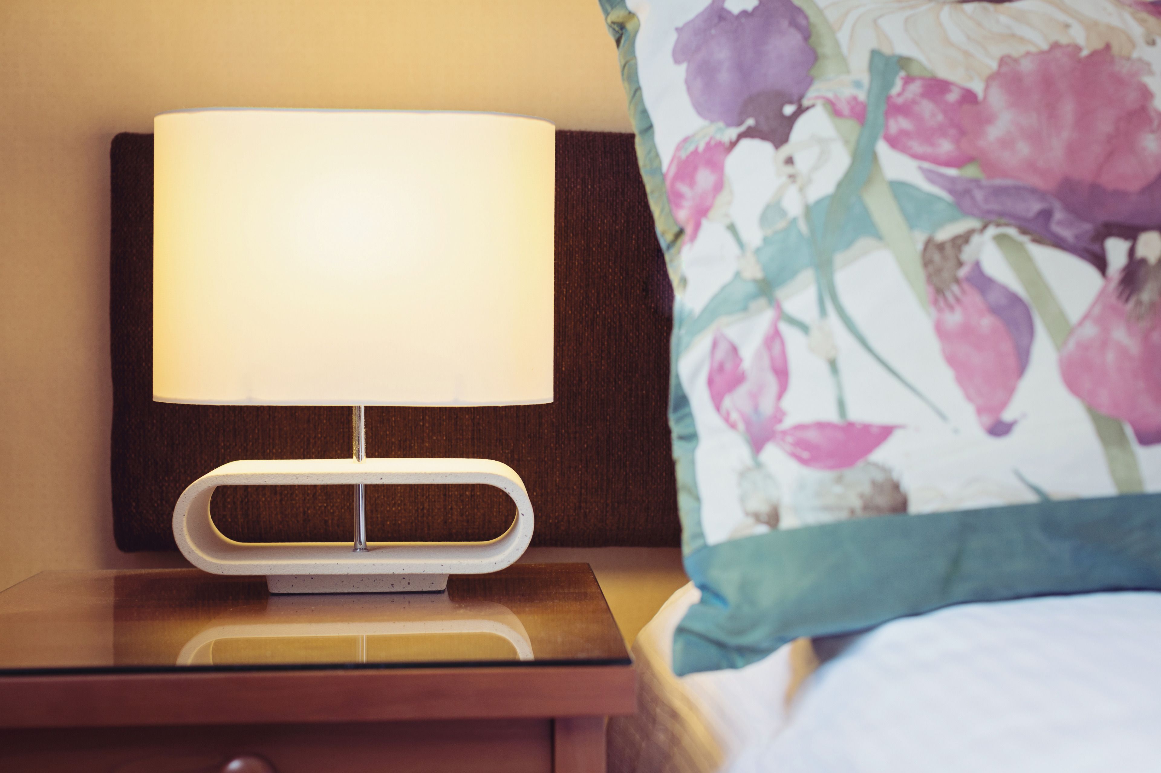 A bedside table with a lamp, a floral pillow, and a wooden headboard.