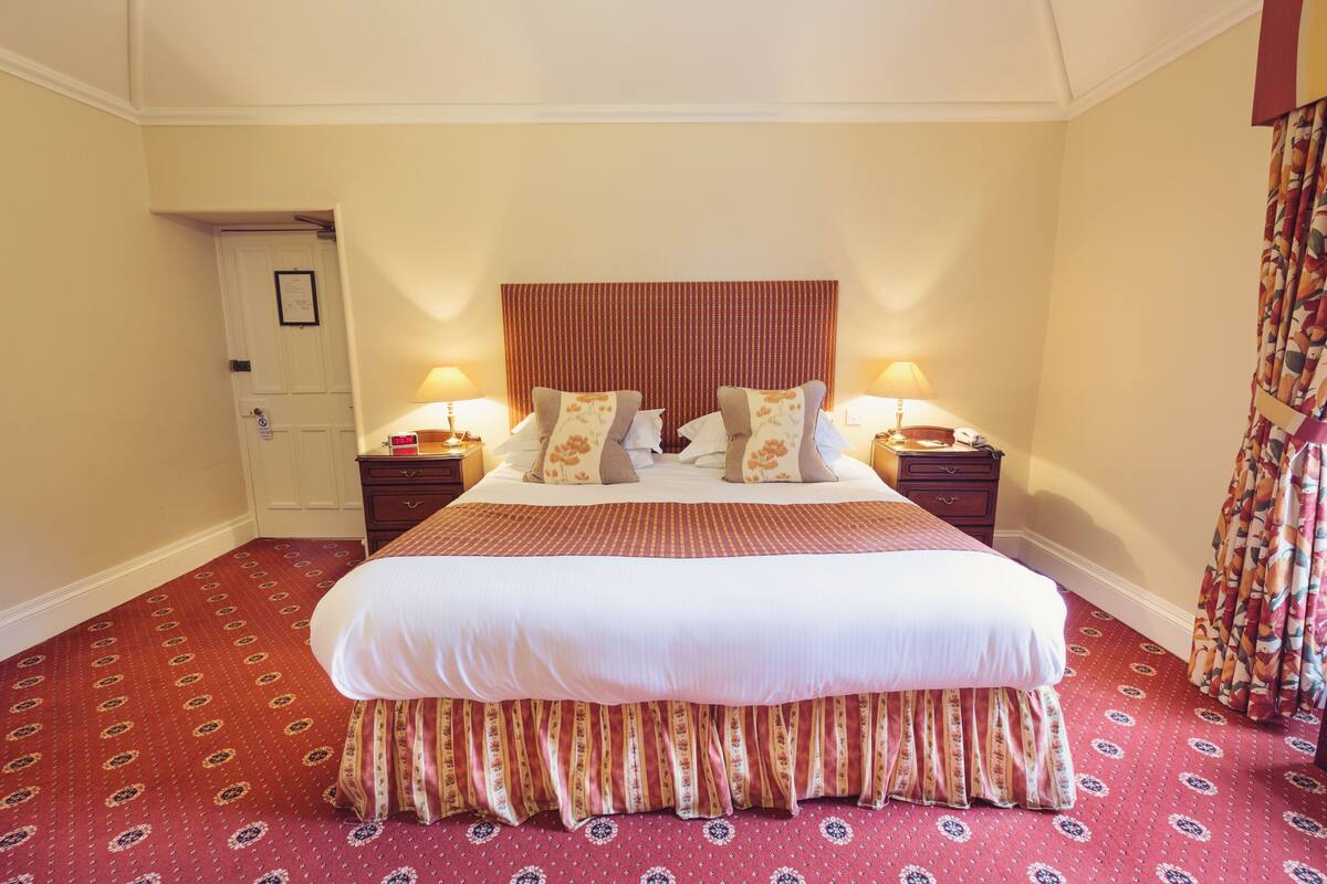 Een hotelkamer met een groot bed, twee nachtkastjes met lampen, een hoofdeinde en een kamerbreed tapijt met een patroon.