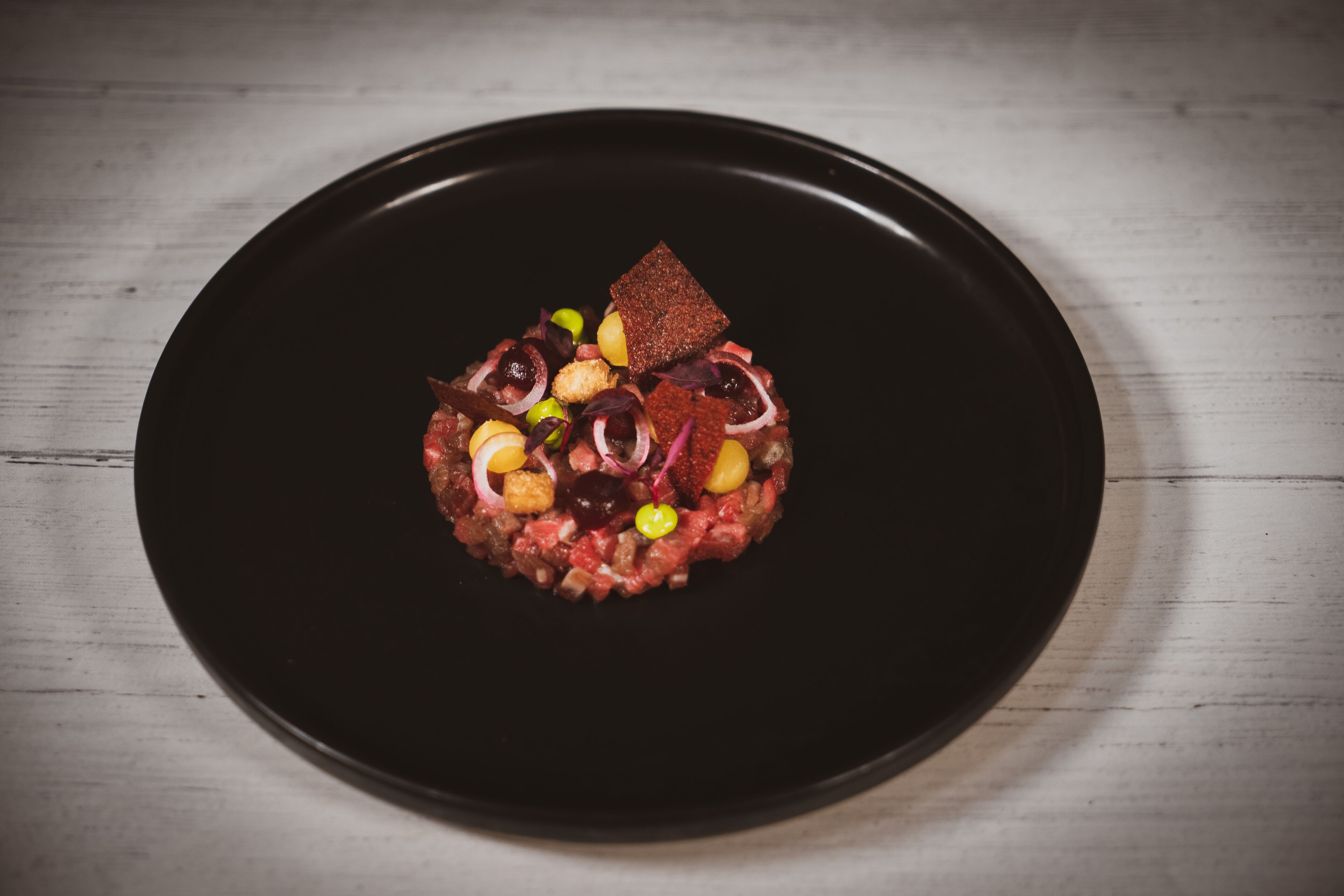 A black plate with a dish of raw meat and various garnishes on a wooden surface.
