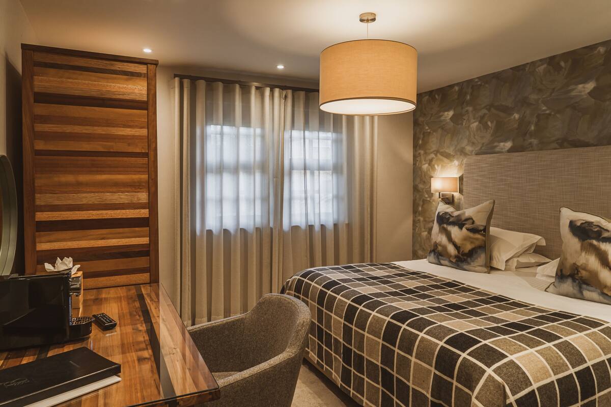 Een hotelkamer met een houten bureau, een stoel, een bed met ruitbeddengoed en een groot raam met gordijnen.