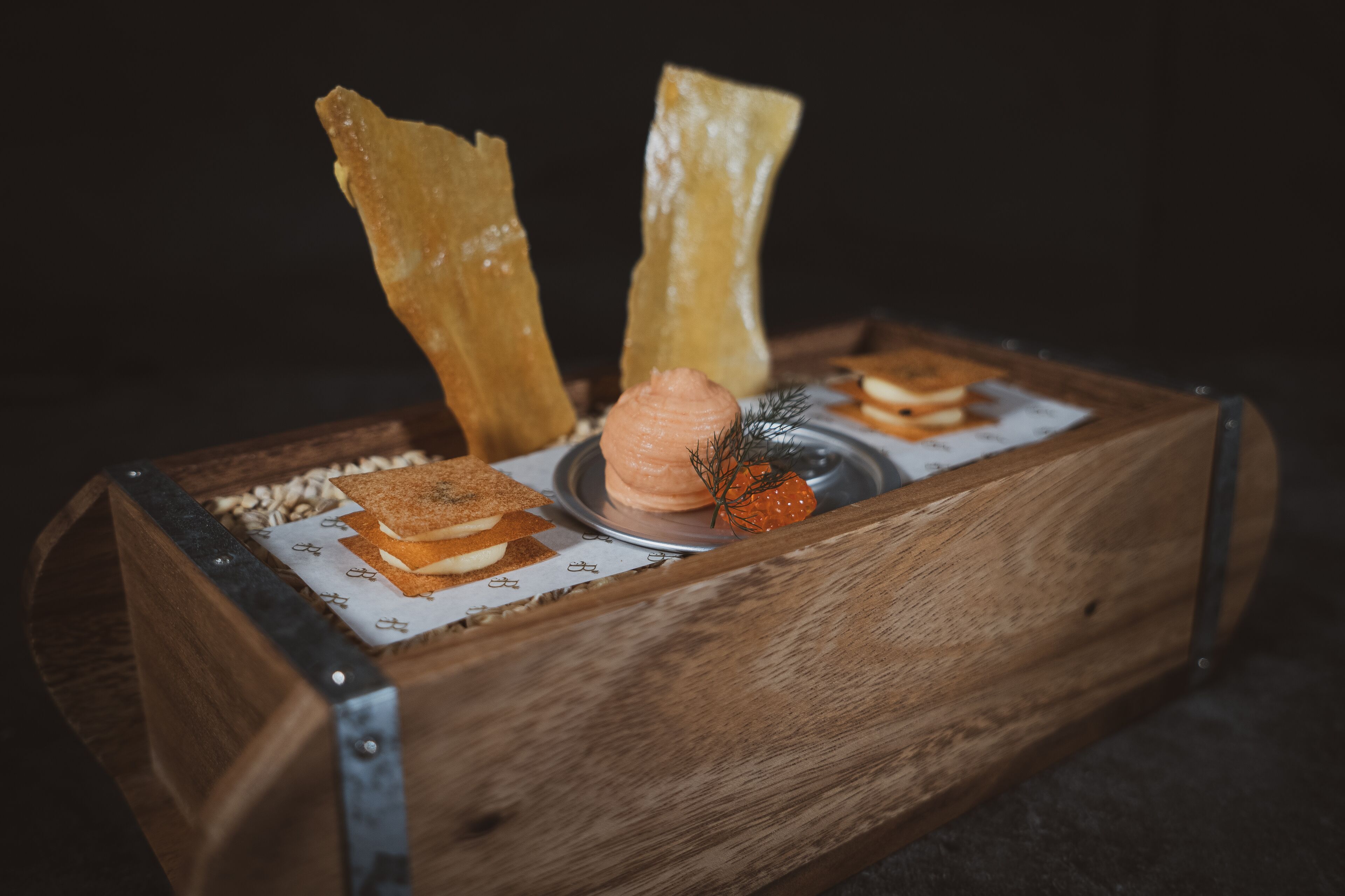 A wooden box with a metal clasp, containing a dessert with a dollop of cream, a sprig of dill, and a small orange fruit.