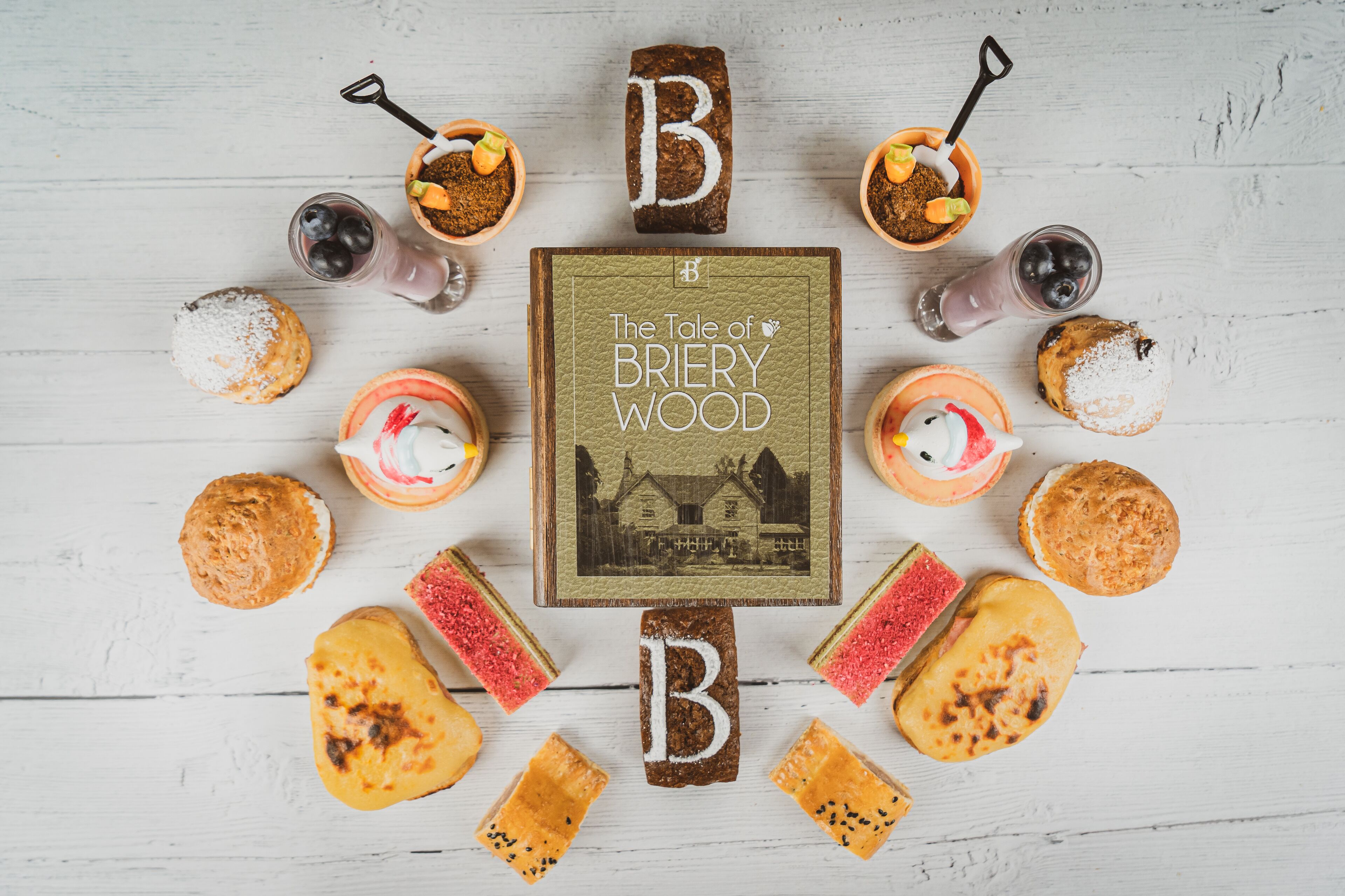 A wooden table with a book titled "The Tale of Briery Wood" surrounded by various pastries and drinks.