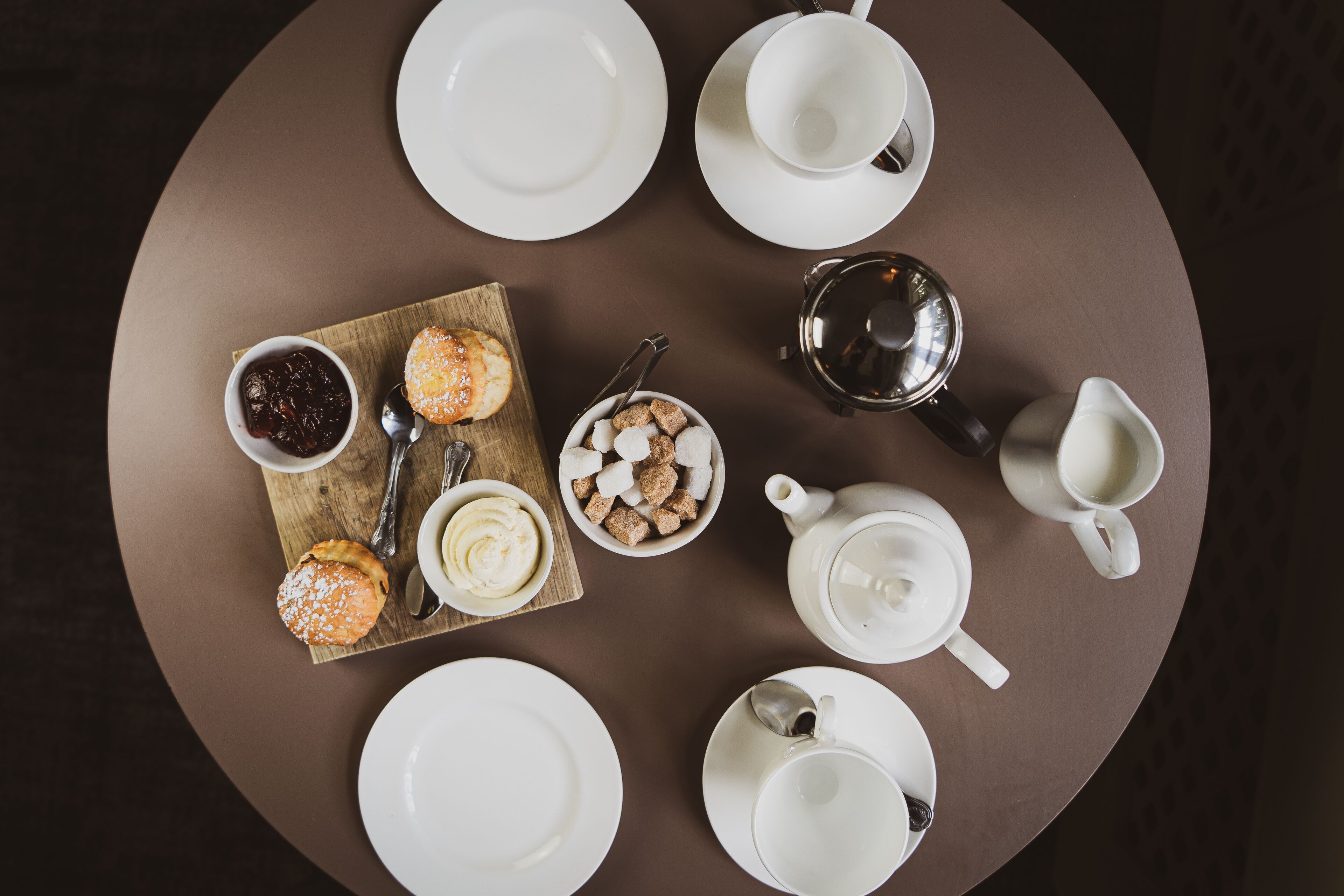 Una mesa redonda con platos blancos, tazas, una tetera, una jarrón, un cuenco de galletas, un recipiente con mermelada y una tabla de madera con pasteles.