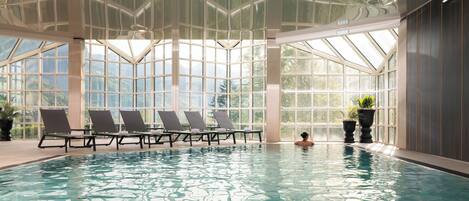 An indoor swimming pool with clear glass walls, lounge chairs, and potted plants.