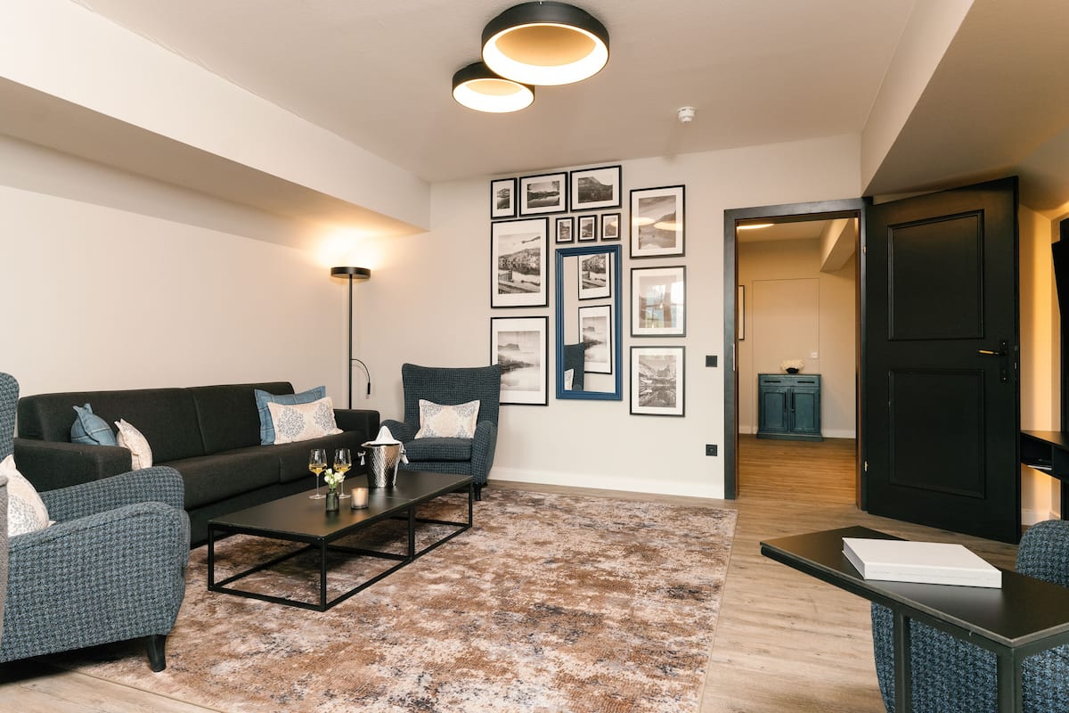 A modern living room with a sofa, armchairs, a coffee table, and a rug. There are framed pictures on the wall and a door leading to another room.