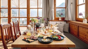 A breakfast table with various foods and drinks, wooden chairs, and large windows with curtains. - MONDI Hotel am Grundlsee (Grundlsee)
