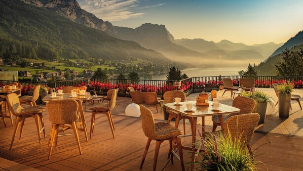 An outdoor terrace with wicker chairs and tables, overlooking a mountainous landscape and a lake. - MONDI Hotel am Grundlsee (Grundlsee)