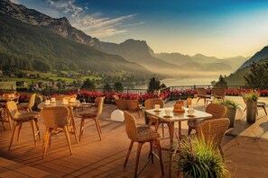 Een terras buiten met rieten stoelen en tafels, met uitzicht op een bergachtig landschap en een meer.