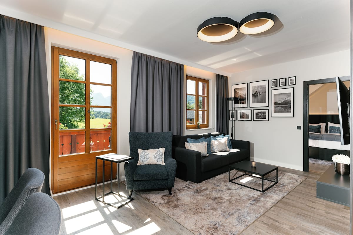 A modern living room with a sofa, armchair, and coffee table, large windows with curtains, and a wall with framed pictures.