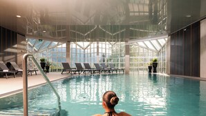 An indoor swimming pool with clear glass walls, lounge chairs, and potted plants. - MONDI Hotel am Grundlsee (Grundlsee)