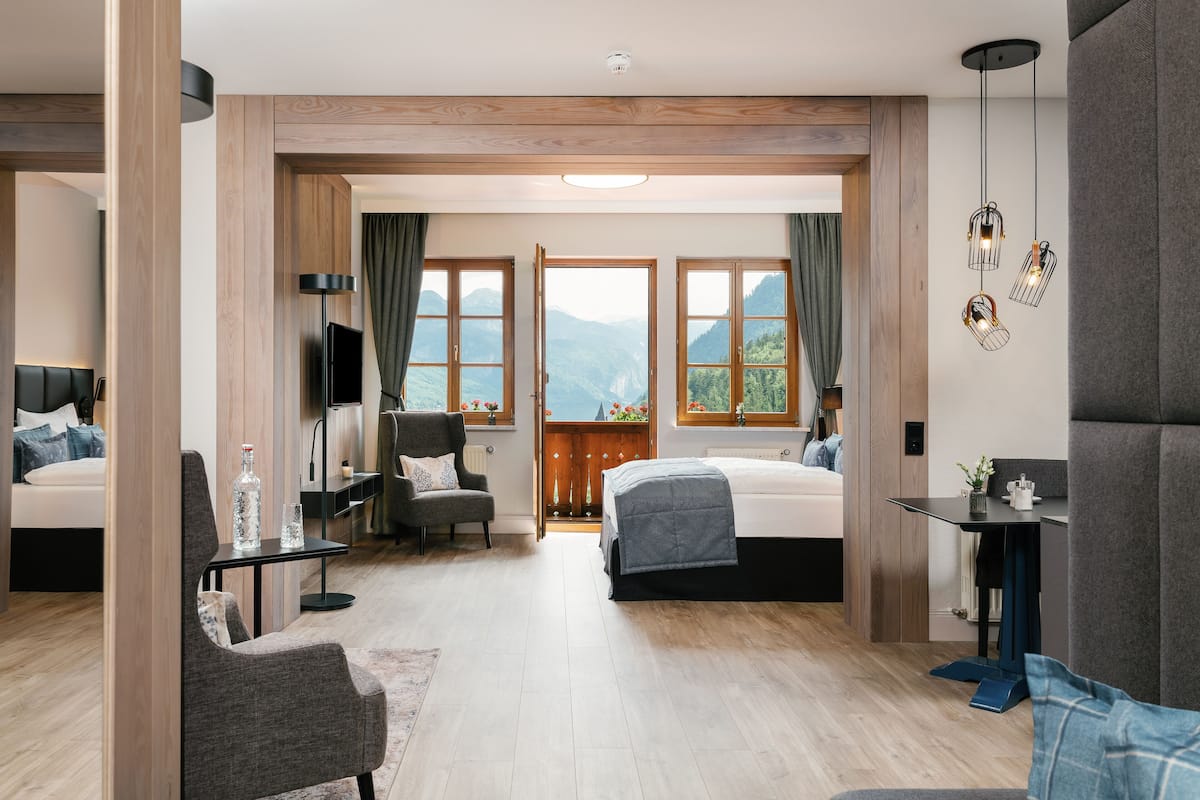 A modern hotel room with two beds, a sitting area with a chair and table, a TV, and large windows with a mountain view.