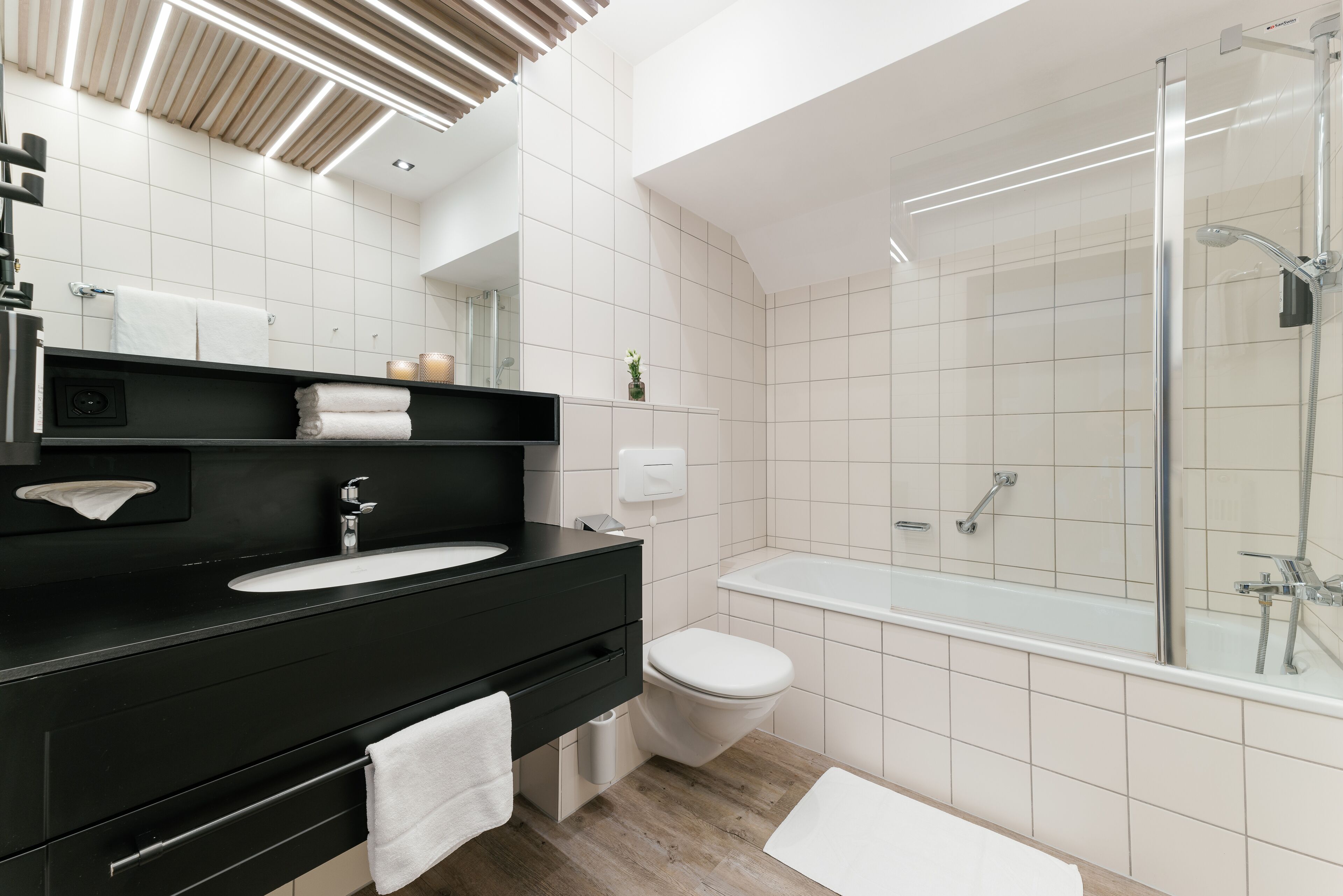 Un baño moderno con un tocador negro, lavamanos blanco y zona de ducha con cerramiento de vidrio.