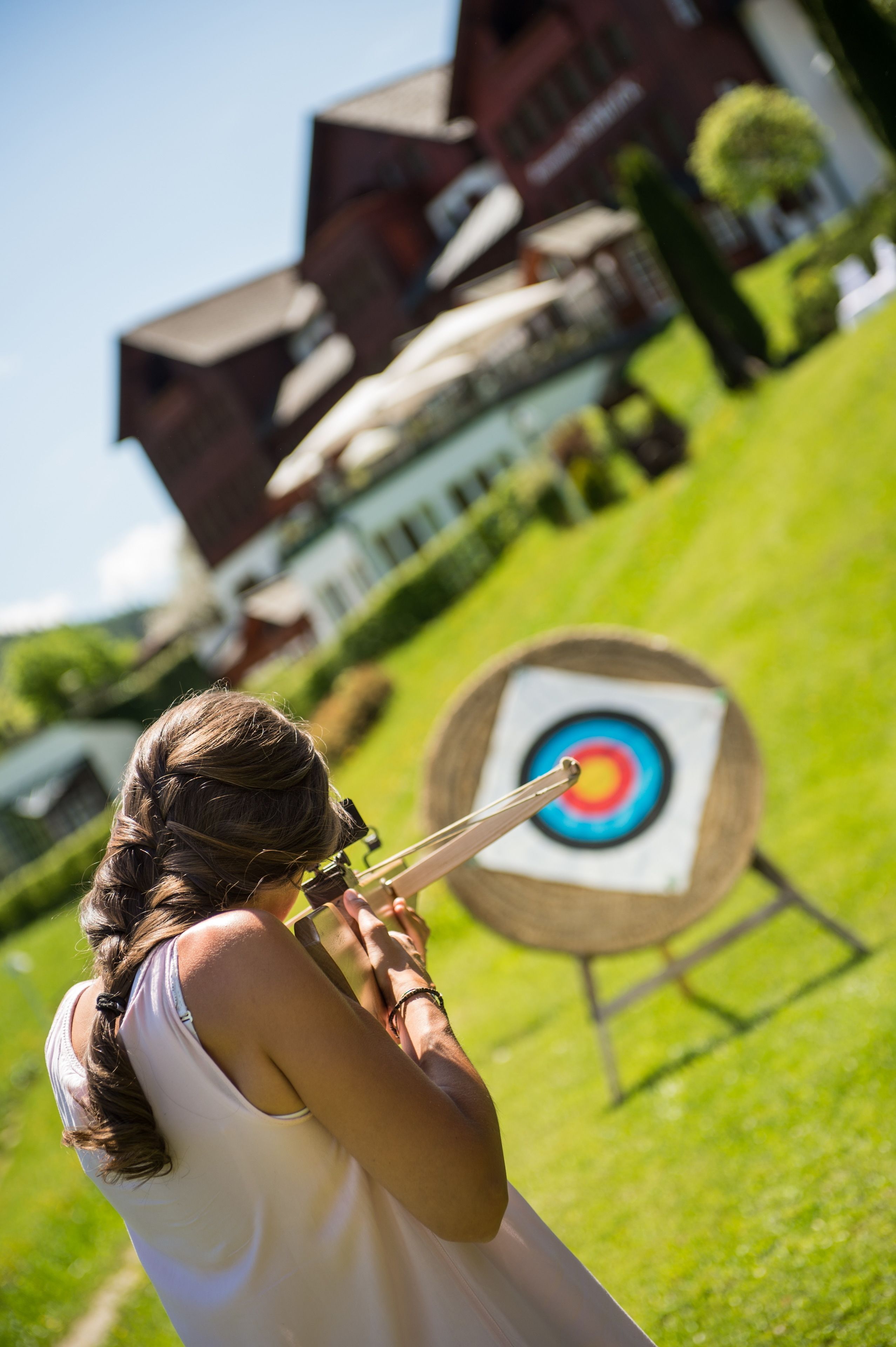 A person aiming an arrow at a target in an outdoor setting with a building in the background.