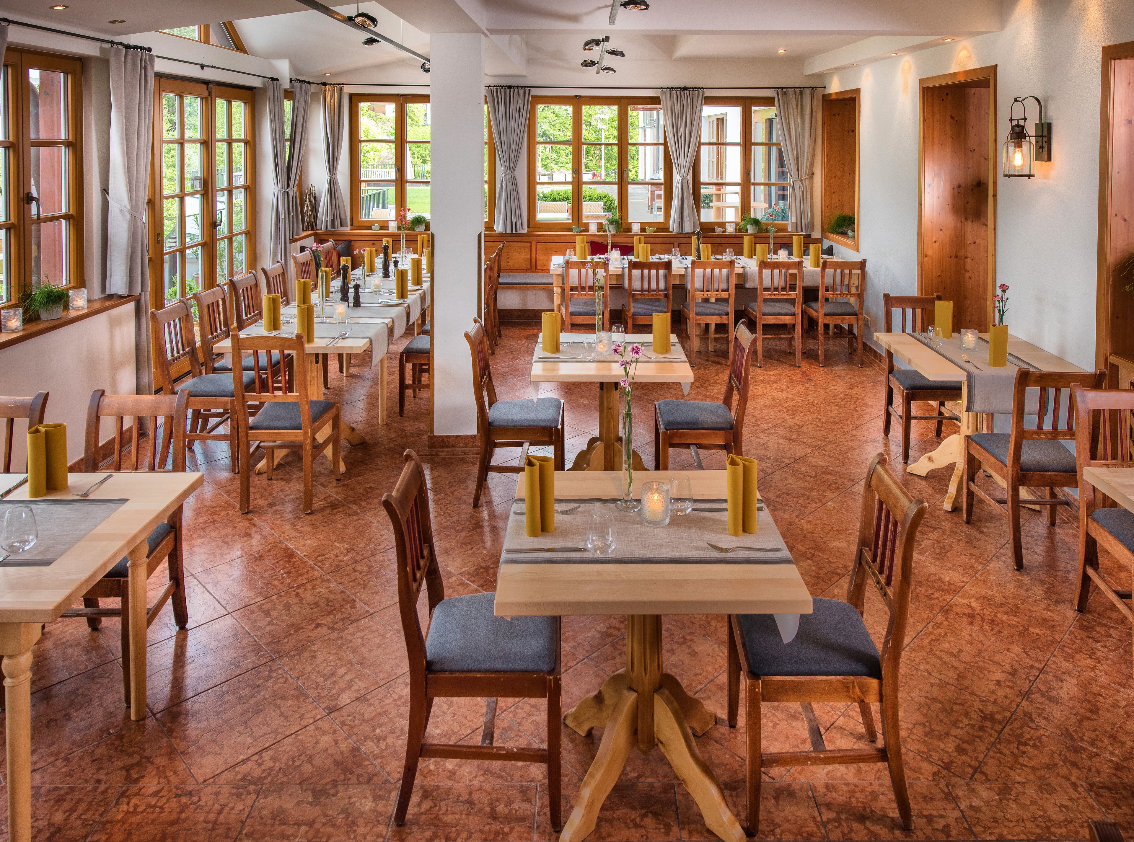 Un restaurante con varias mesas y sillas, ventanas amplias con cortinas y suelo de baldosas.