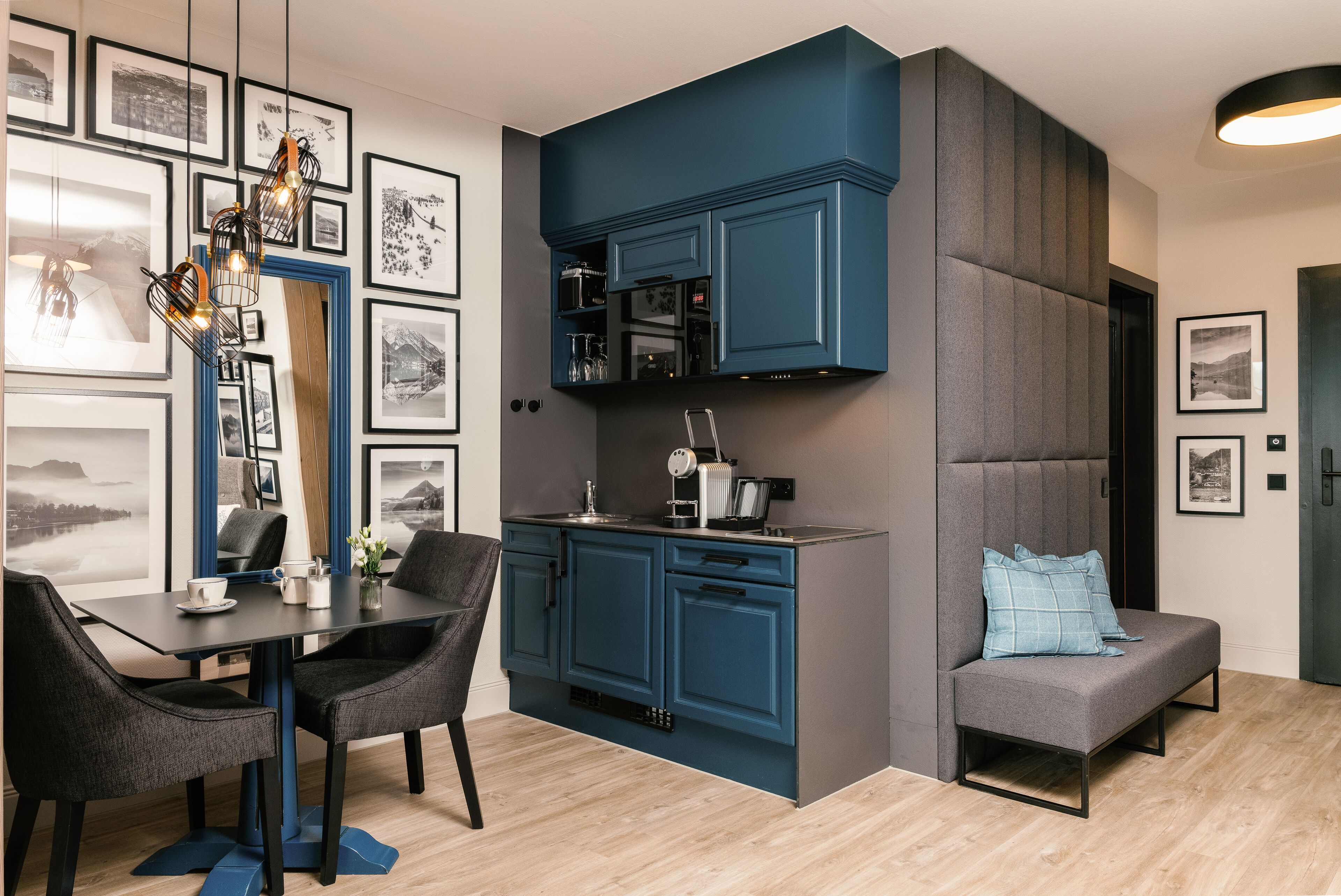 Eine moderne Küche mit blauen Schränken, ein Essbereich mit Tisch und Stühlen sowie ein graues Sofa mit einem blauen Kissen.