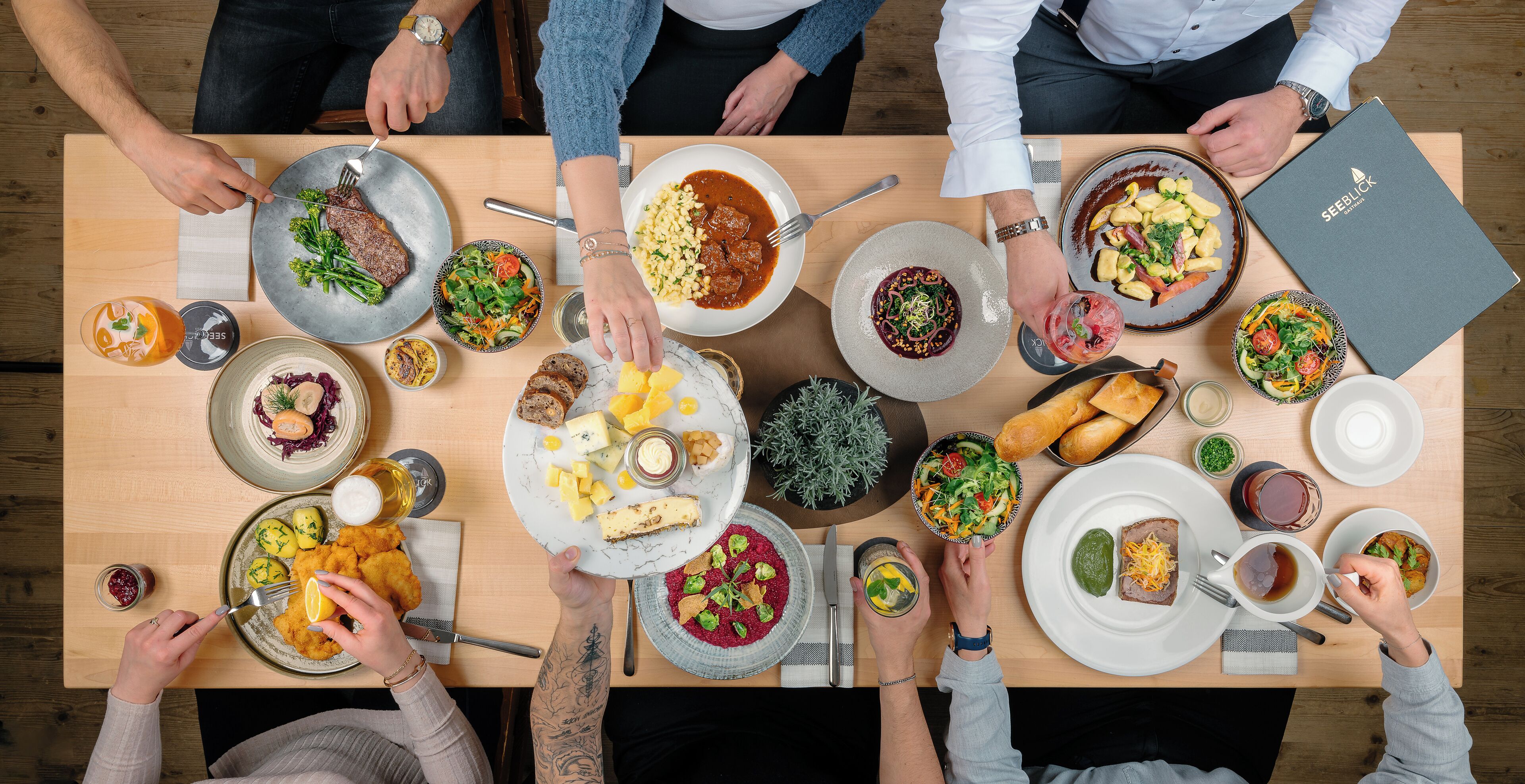 A group of people dining at a wooden table with various dishes, including salads, meats, and bread.