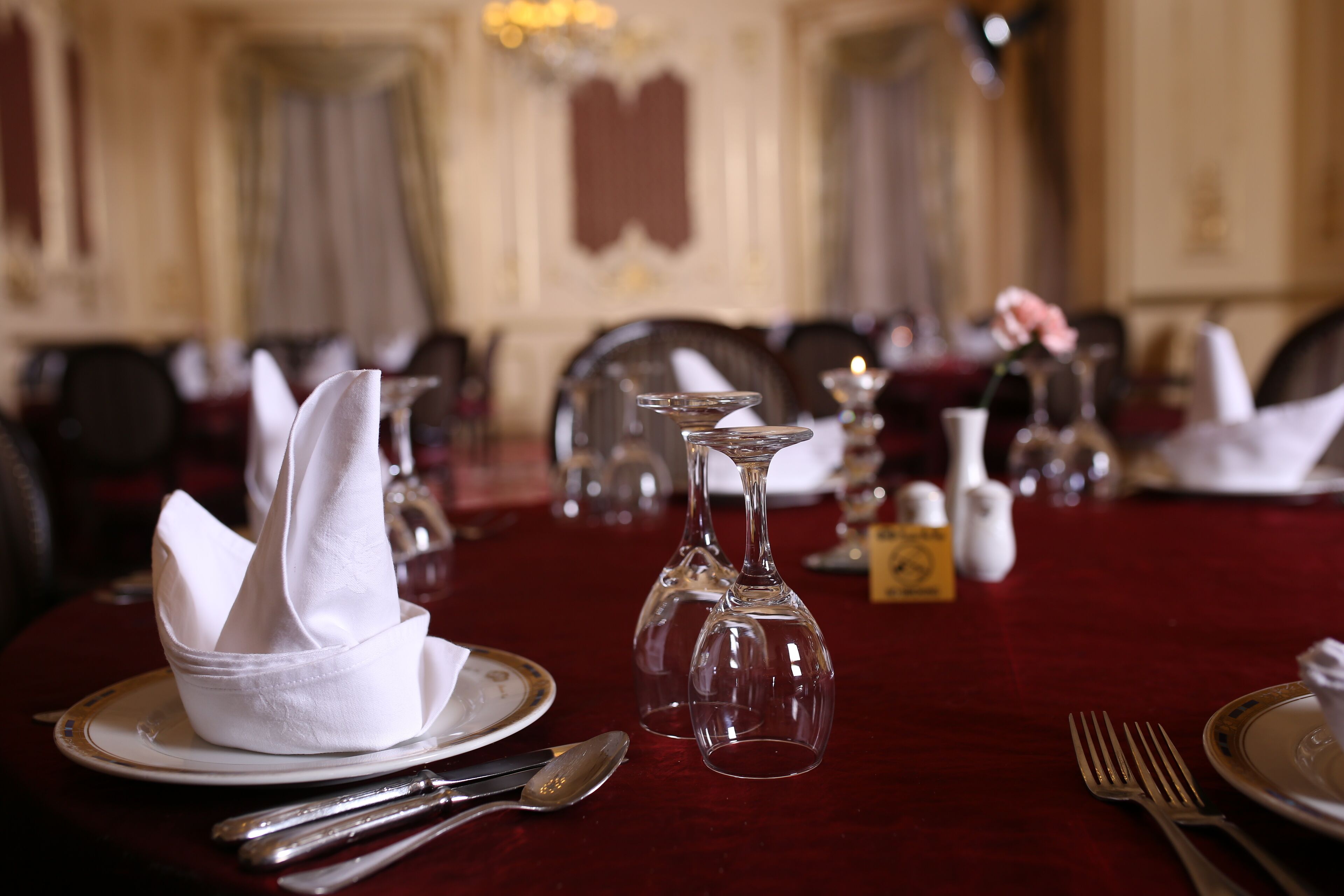 A dining table set with white napkins, silverware, and clear wine glasses, with a red tablecloth and a chandelier in the background.