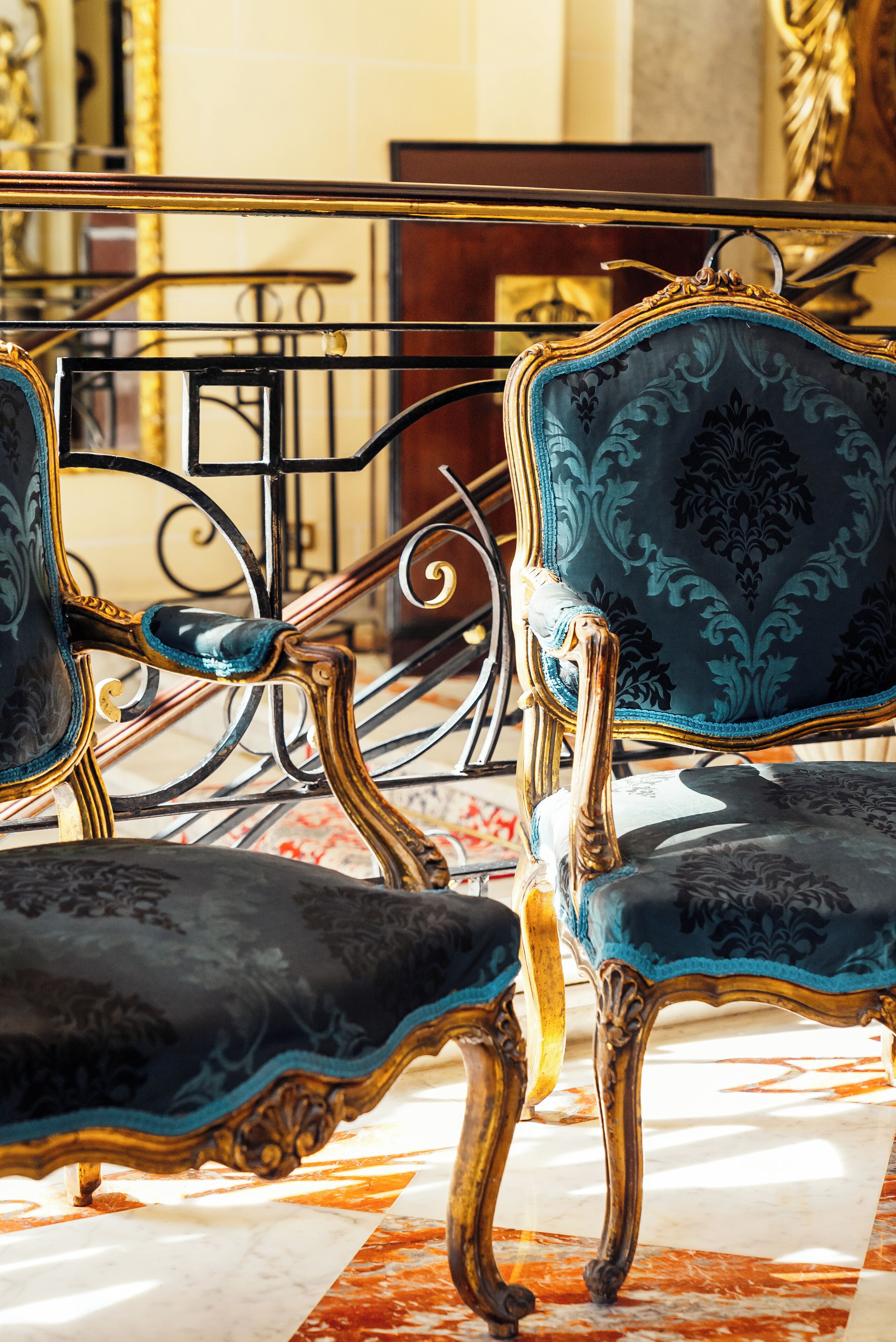 Sillones ornamentados con adornos dorados y tapicería azul oscuro en una habitación luminosa con suelo de mármol.