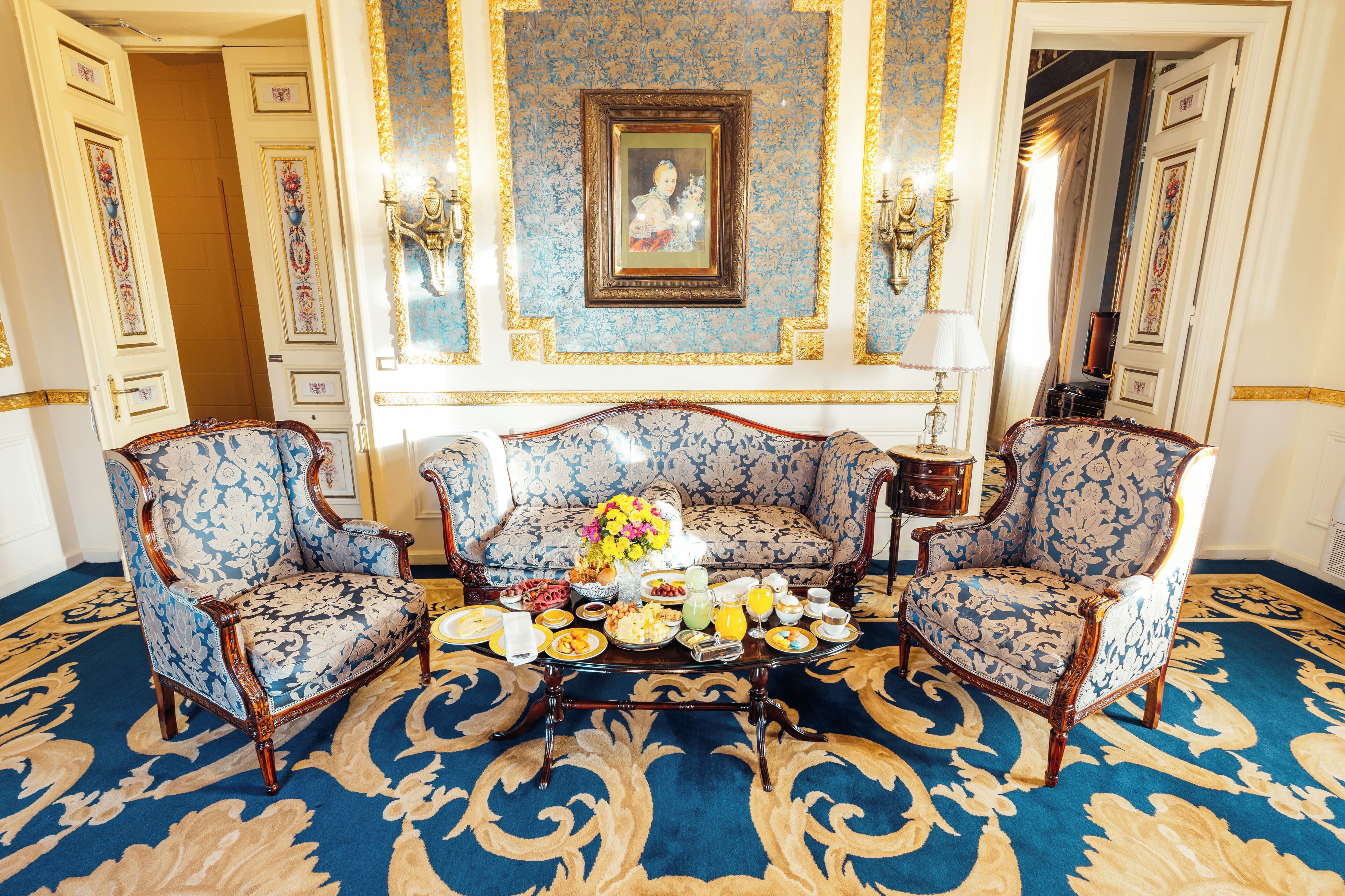 A room with a patterned carpet, a sofa, two armchairs, a coffee table with breakfast items, and a framed picture on the wall.