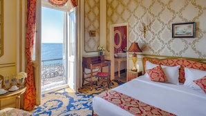 A hotel room with a bed, a desk, a chair, a nightstand, and a view of the sea.
