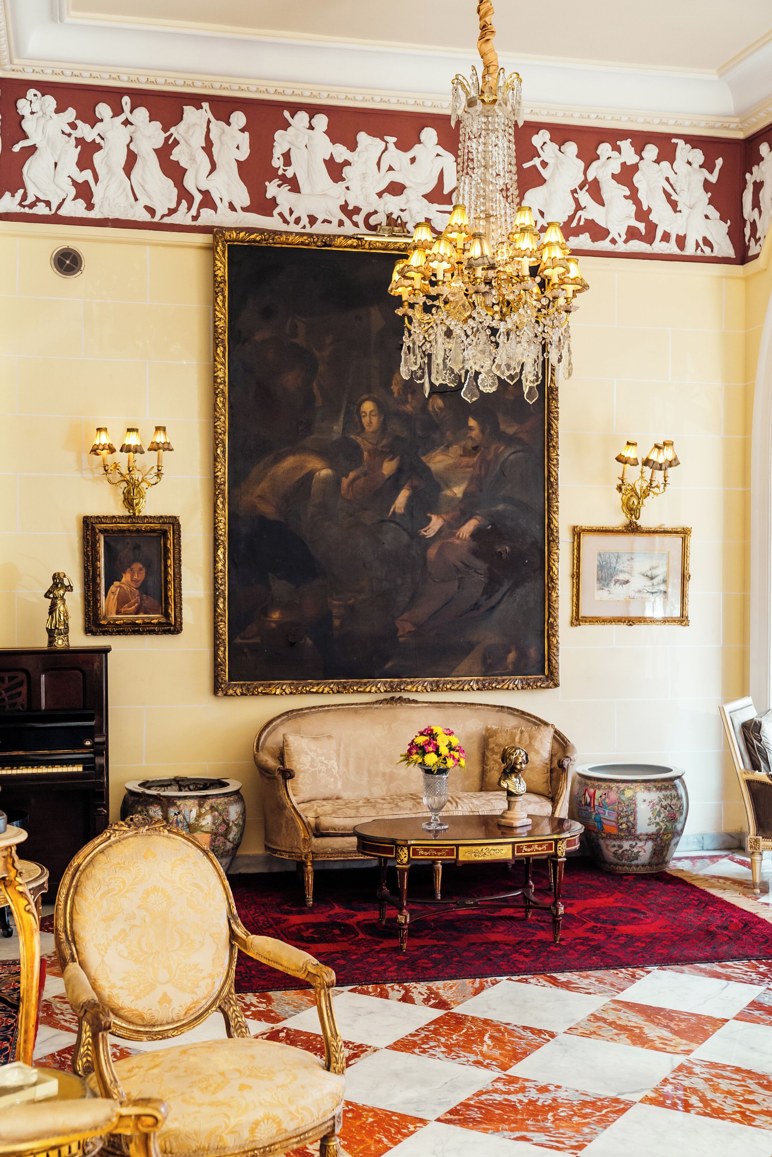 A grand room with a large classical painting, ornate furniture, and a chandelier.