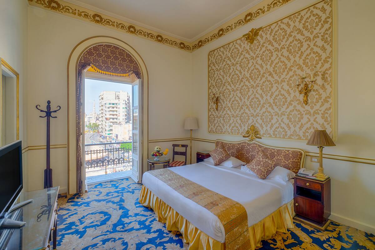 A hotel room with a large bed, a TV, a balcony with a view of buildings, and a patterned wall.