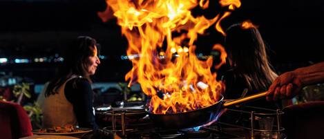 A kitchen with a pan on a stove, flames, and a person cooking.