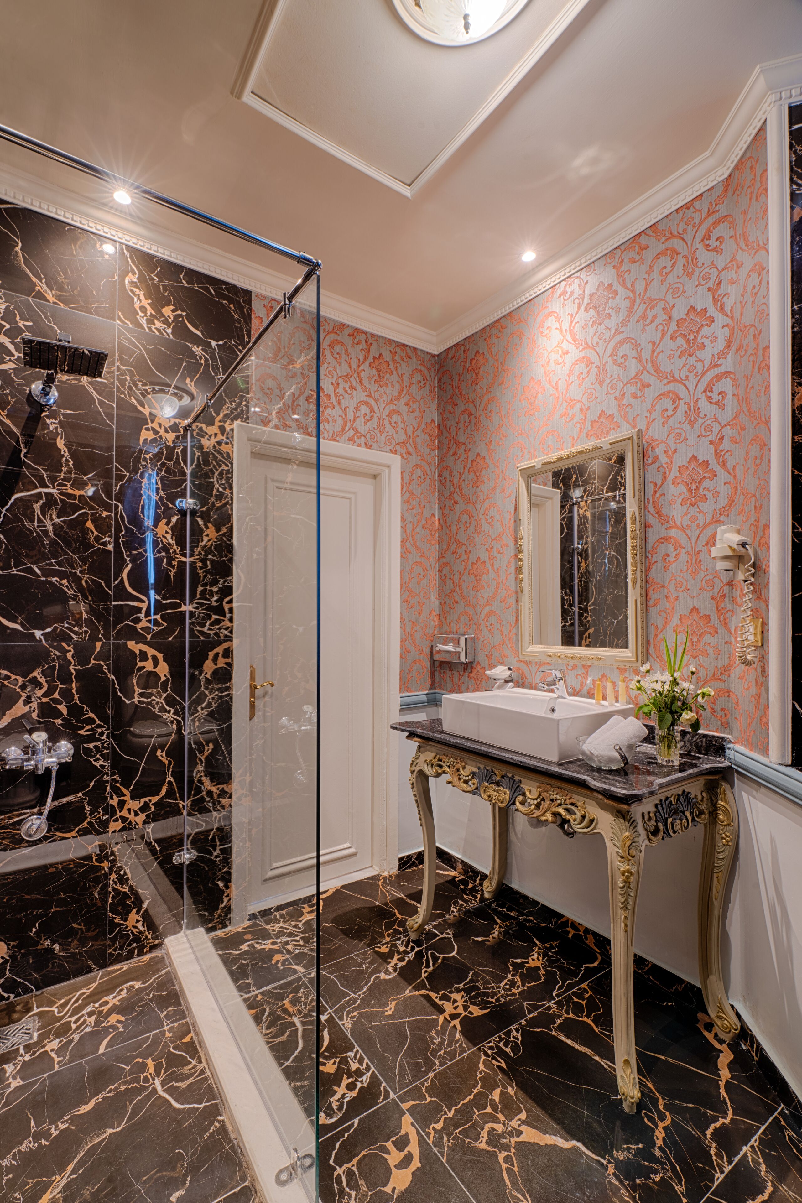 Un baño con ducha de vidrio, suelo de mármol, lavamanos con espejo y decoraciones de pared ornamentales.