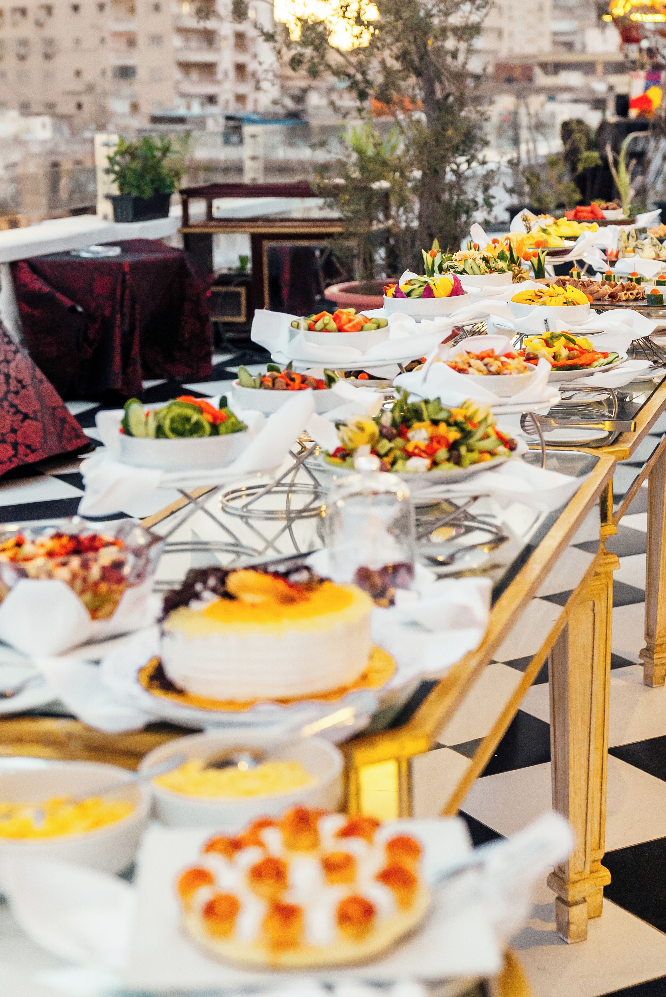 Un tavolo da buffet con diversi piatti e una torta, su un pavimento a scacchi con uno skyline urbano sullo sfondo.
