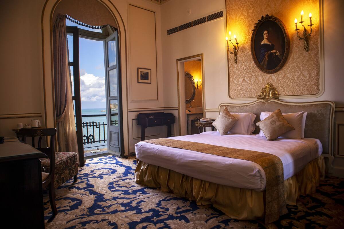 A hotel room with a large bed, a view of the sea through an open door, a chandelier, and a framed portrait on the wall.