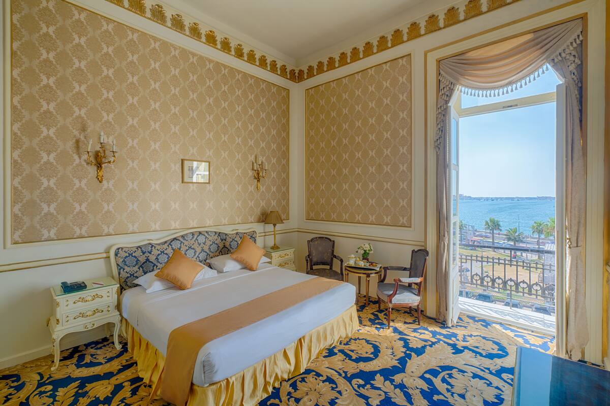 A hotel room with a large bed, a small table, a chair, and a balcony with a view of the sea.