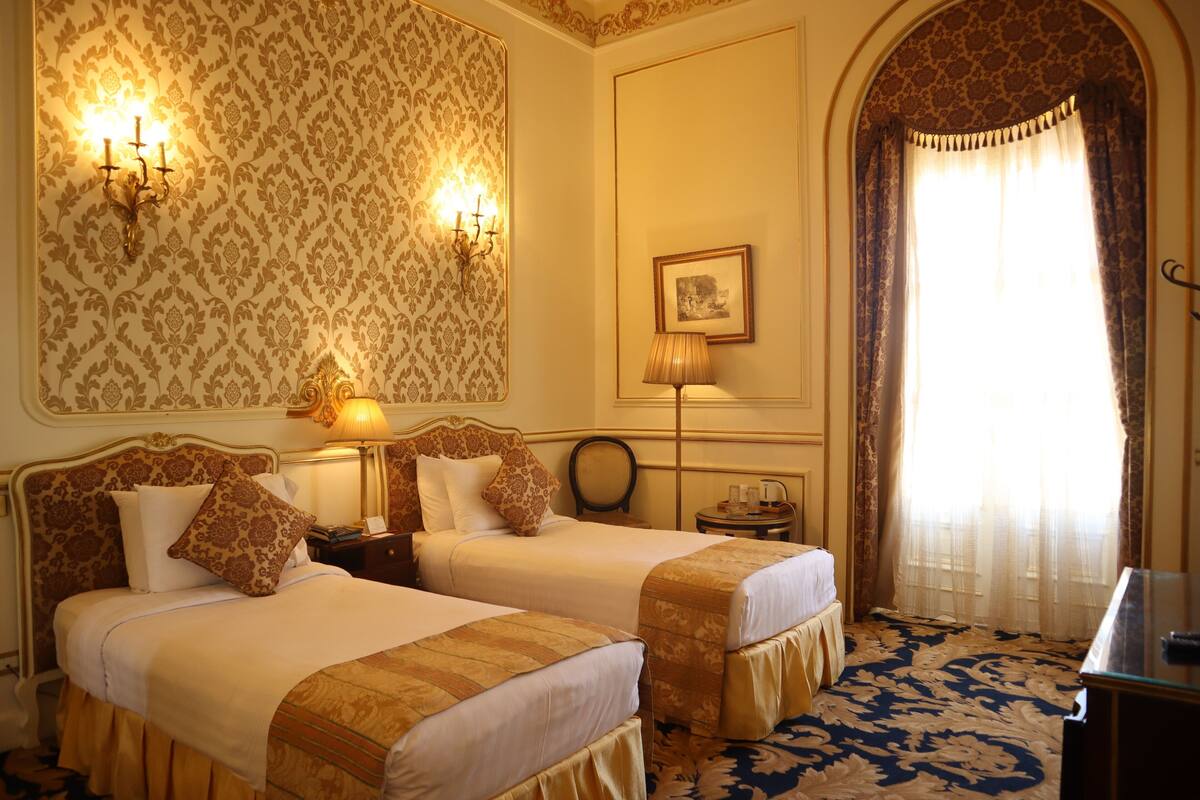 A hotel room with two beds, a lamp, a picture on the wall, and a large window with curtains.