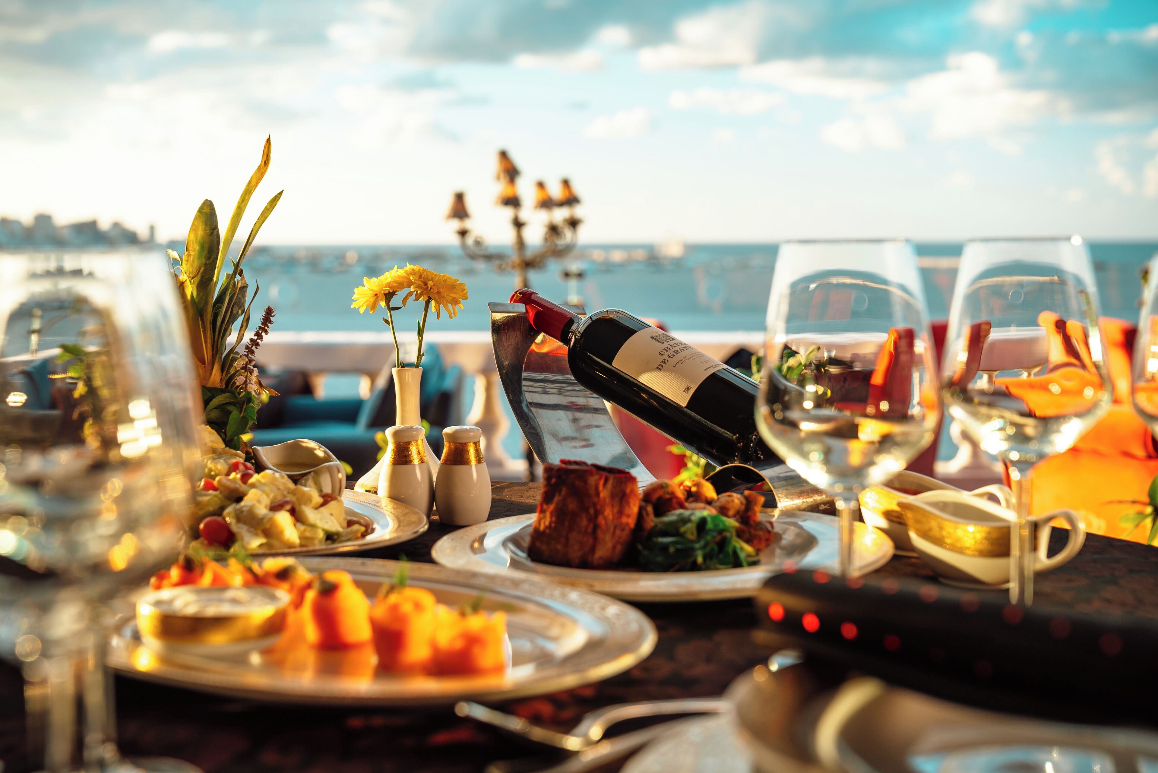 Une table dressée pour un repas, avec une bouteille de vin, des verres et une vue sur la mer.