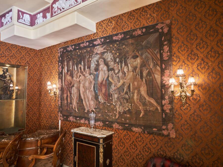 A room with ornate wallpaper, a large classical painting, a marble-topped table, and a statue.