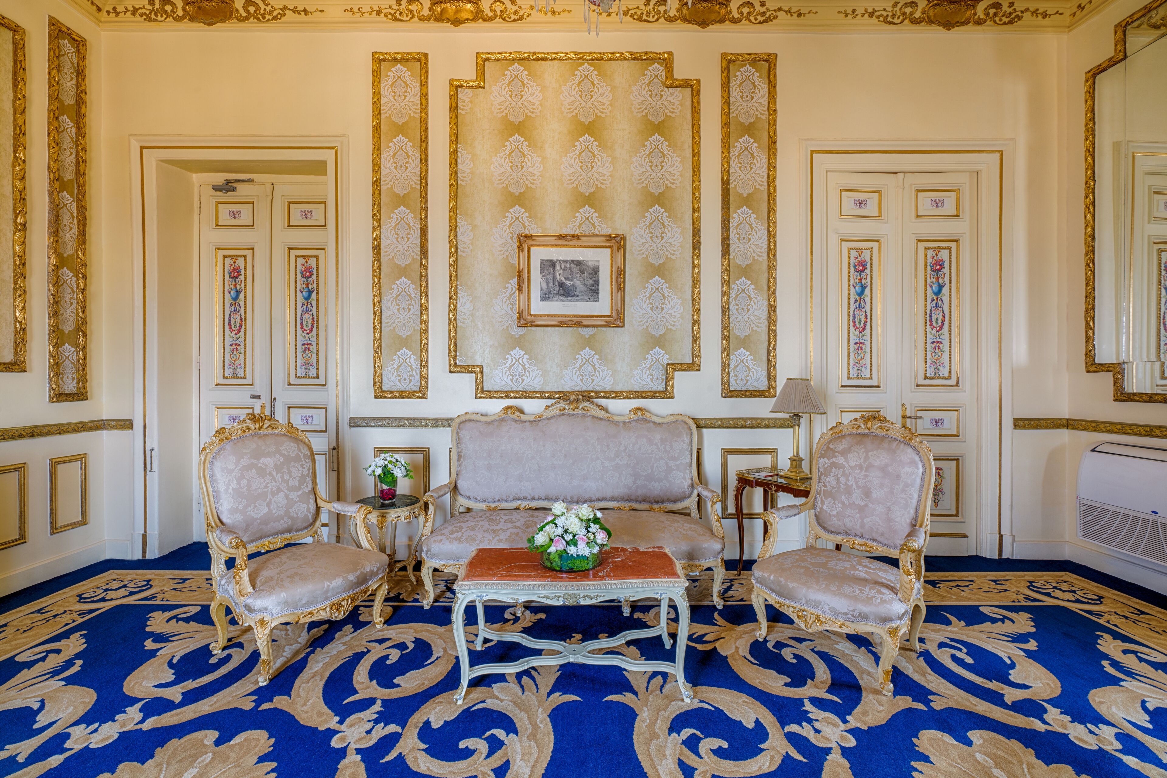 A room with a blue patterned carpet, a sofa, two armchairs, a small table with a vase of flowers, and a framed picture on the wall.