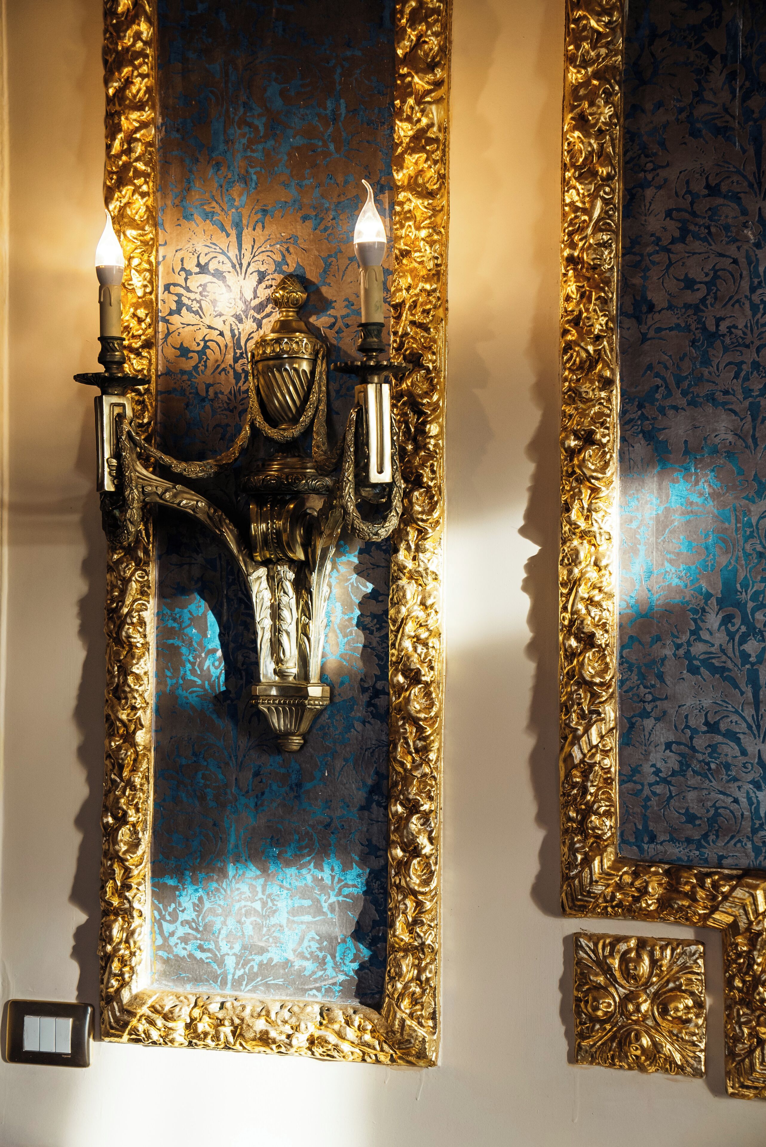 Eine Wandleuchte mit zwei Kerzen, aufwändiger Goldverzierung und einem blauen, gemusterten Hintergrund.