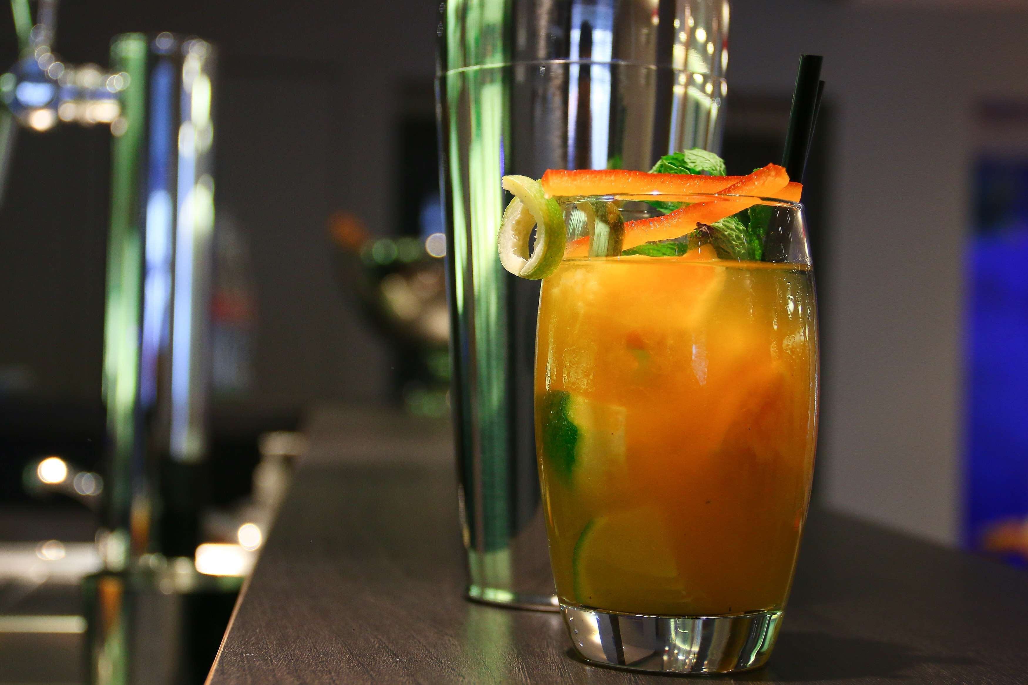 A cocktail with a garnish, a straw, and a bar setting with a shaker in the background.