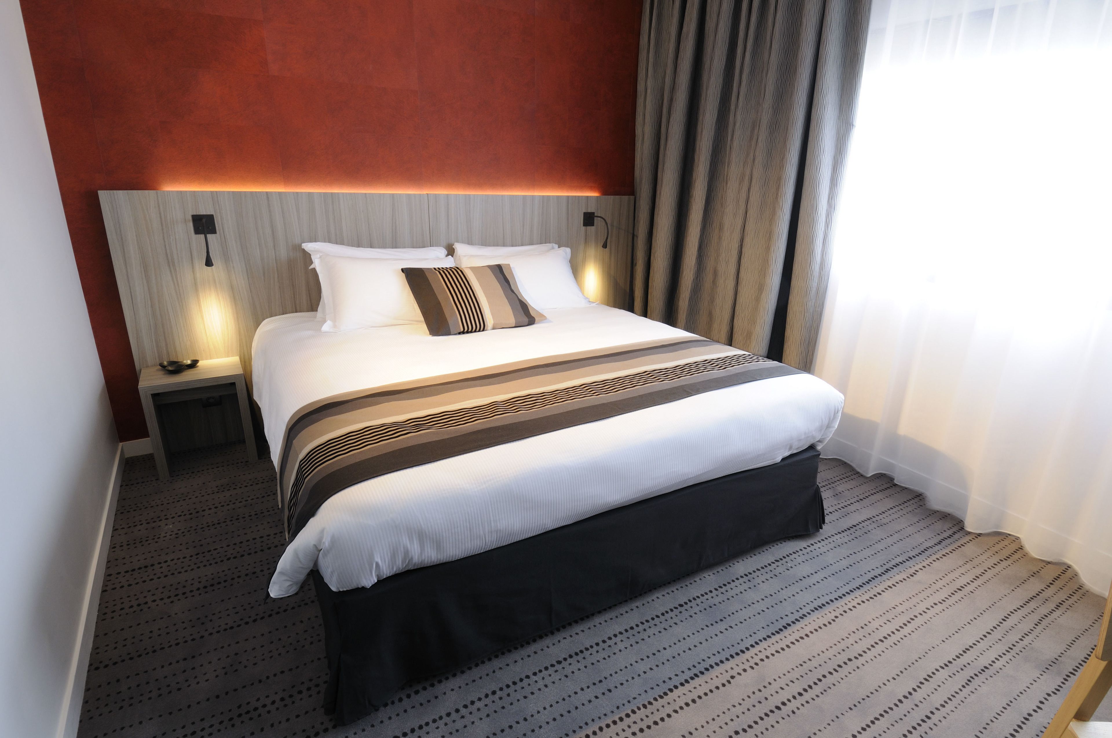 Une chambre d’hôtel avec un grand lit, une table de chevet, une lampe fixée au mur et une fenêtre avec des rideaux.