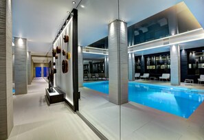 A modern indoor swimming pool with lounge chairs and a glass wall.
