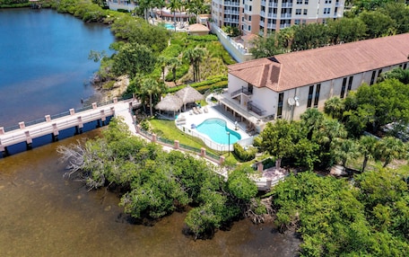 An aerial view of a resort with a swimming pool, lush greenery, and a bridge over water.