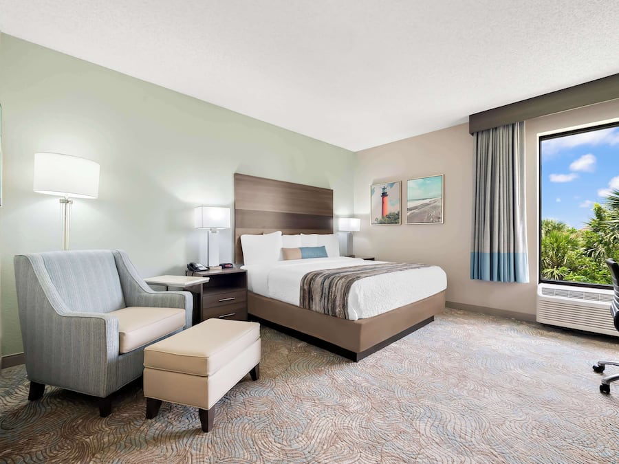 A hotel room with a bed, a desk, a chair, a small table, and a window with a view of greenery.