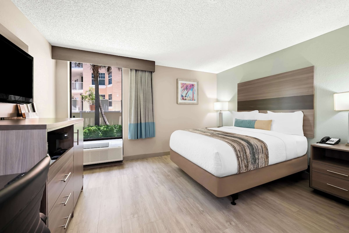 A hotel room with a large bed, a desk with drawers, a window with a view of buildings, and a mounted TV.