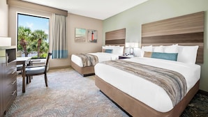 Une chambre d’hôtel avec deux lits, un bureau et une chaise. Il y a une fenêtre donnant sur des palmiers et un immeuble.