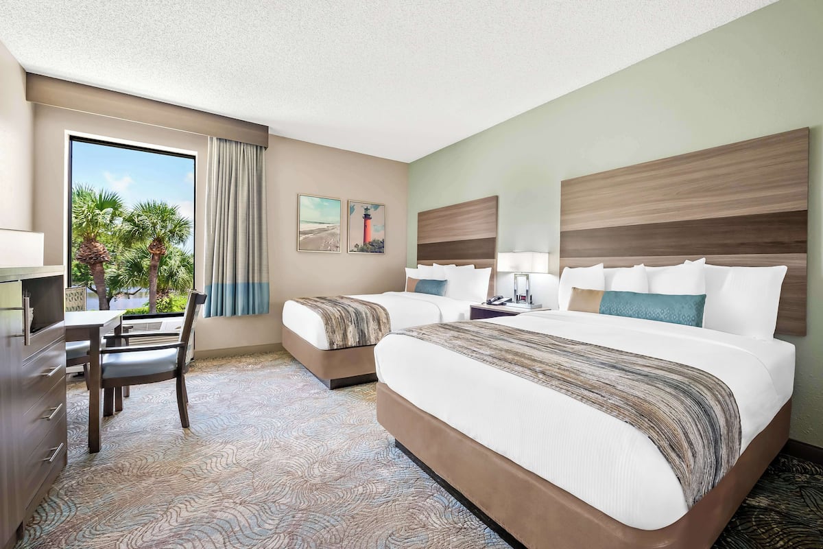 A hotel room with two beds, a desk, and a chair. There is a window with a view of palm trees and a building.