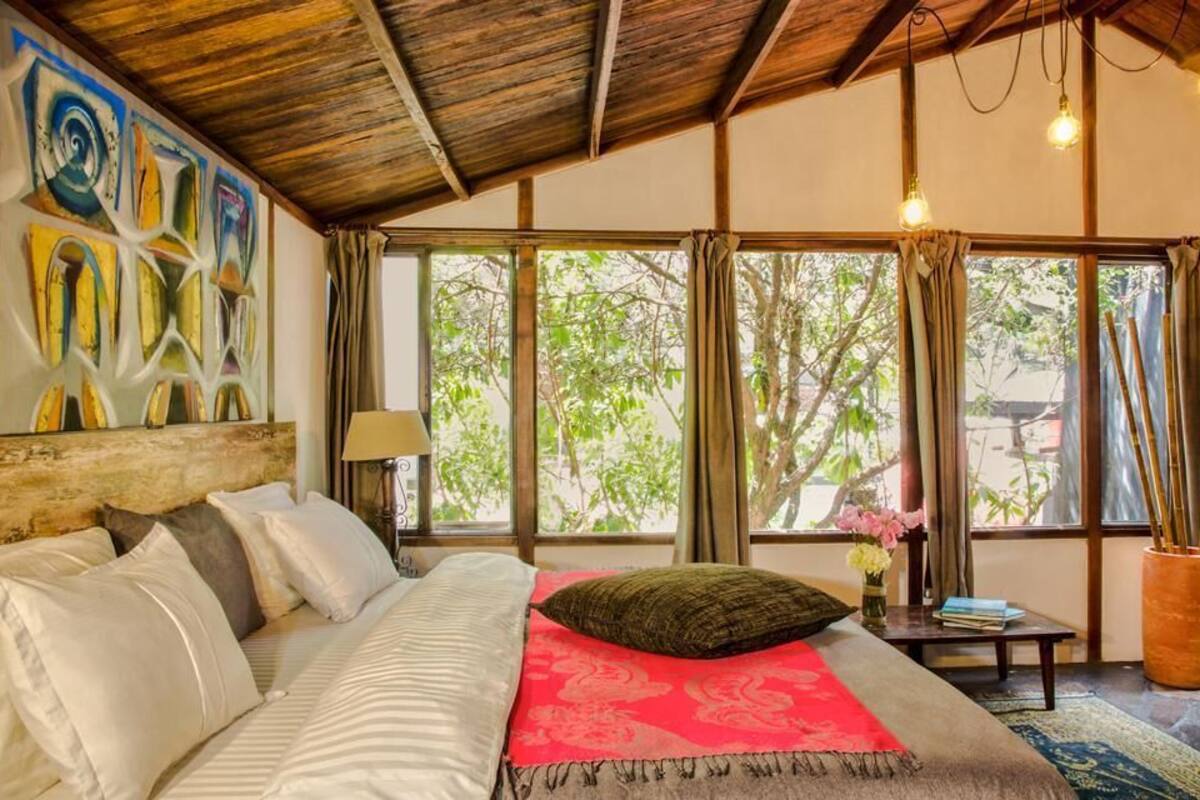 A bedroom with a bed, a nightstand, a lamp, a window with curtains, and a view of trees.