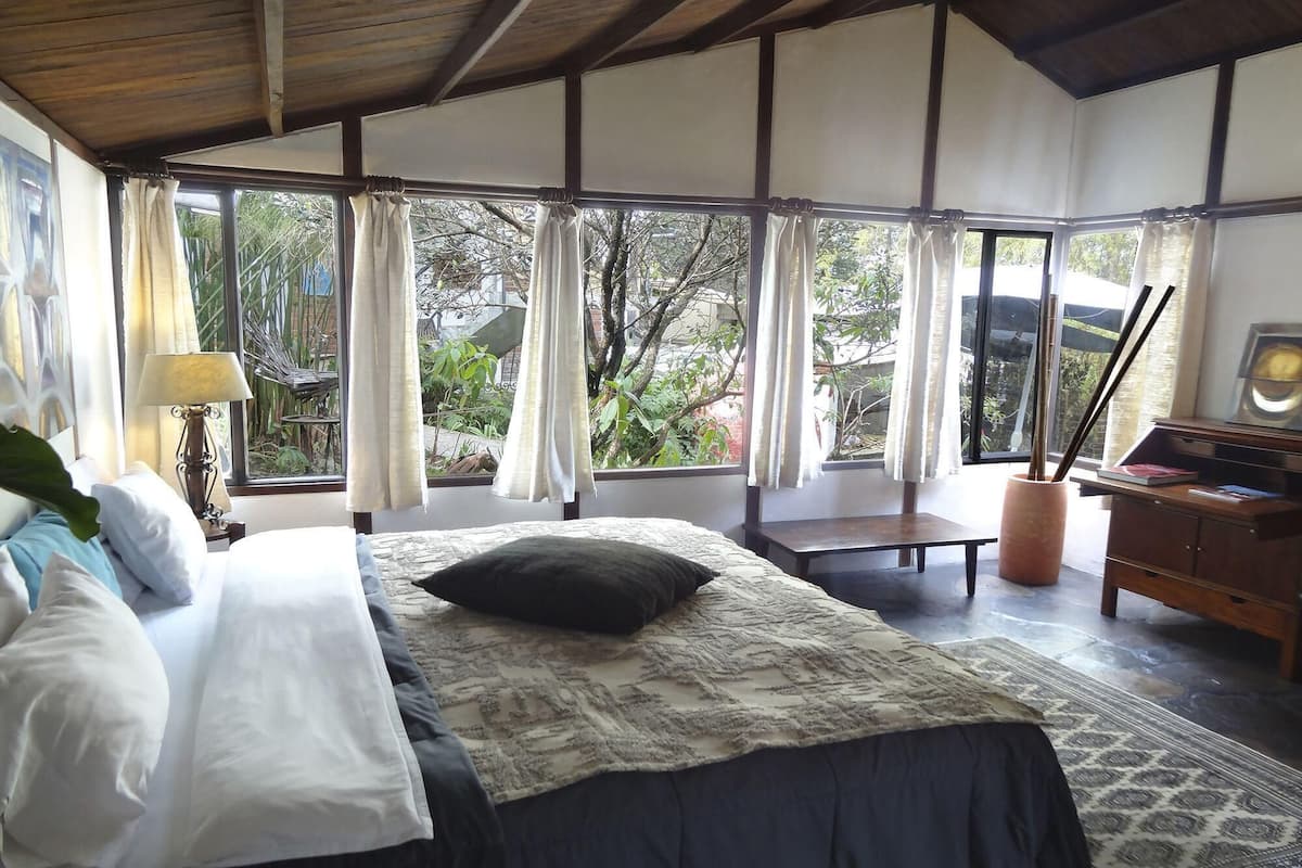 A bedroom with a bed, bedside table, lamp, and a view of greenery through large windows.